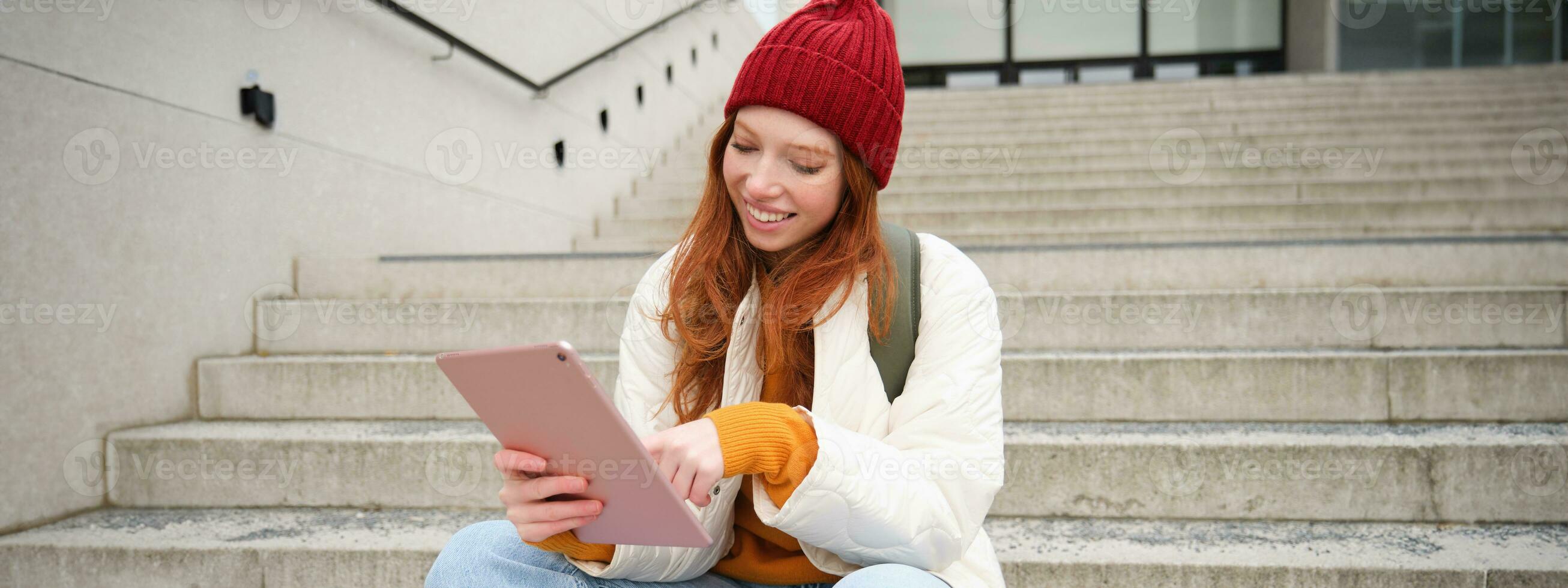 Happy stylish redhead girl, student in red hat, holds digital tablet, uses social media app, searches something online, connects to wifi photo