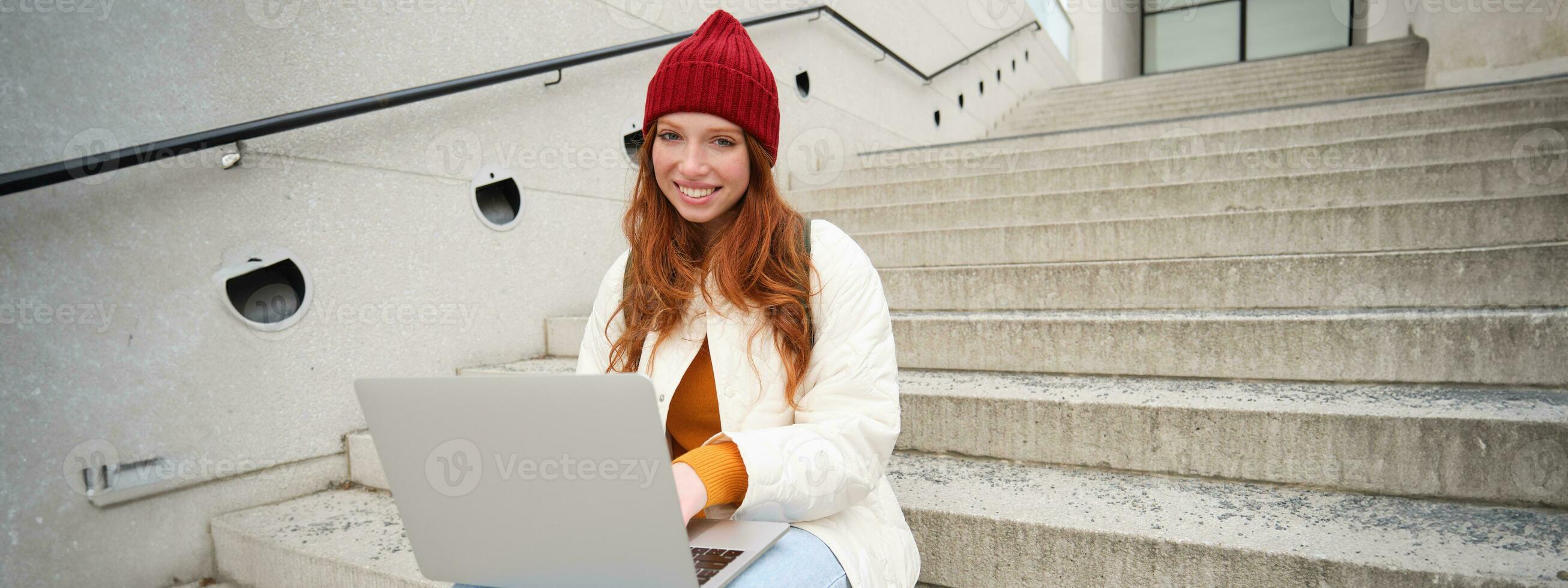 Smiling redhead girl, young woman typing on laptop keyboard, sitting outdoors on stairs with computer, working remote, doing her homework on fresh air photo