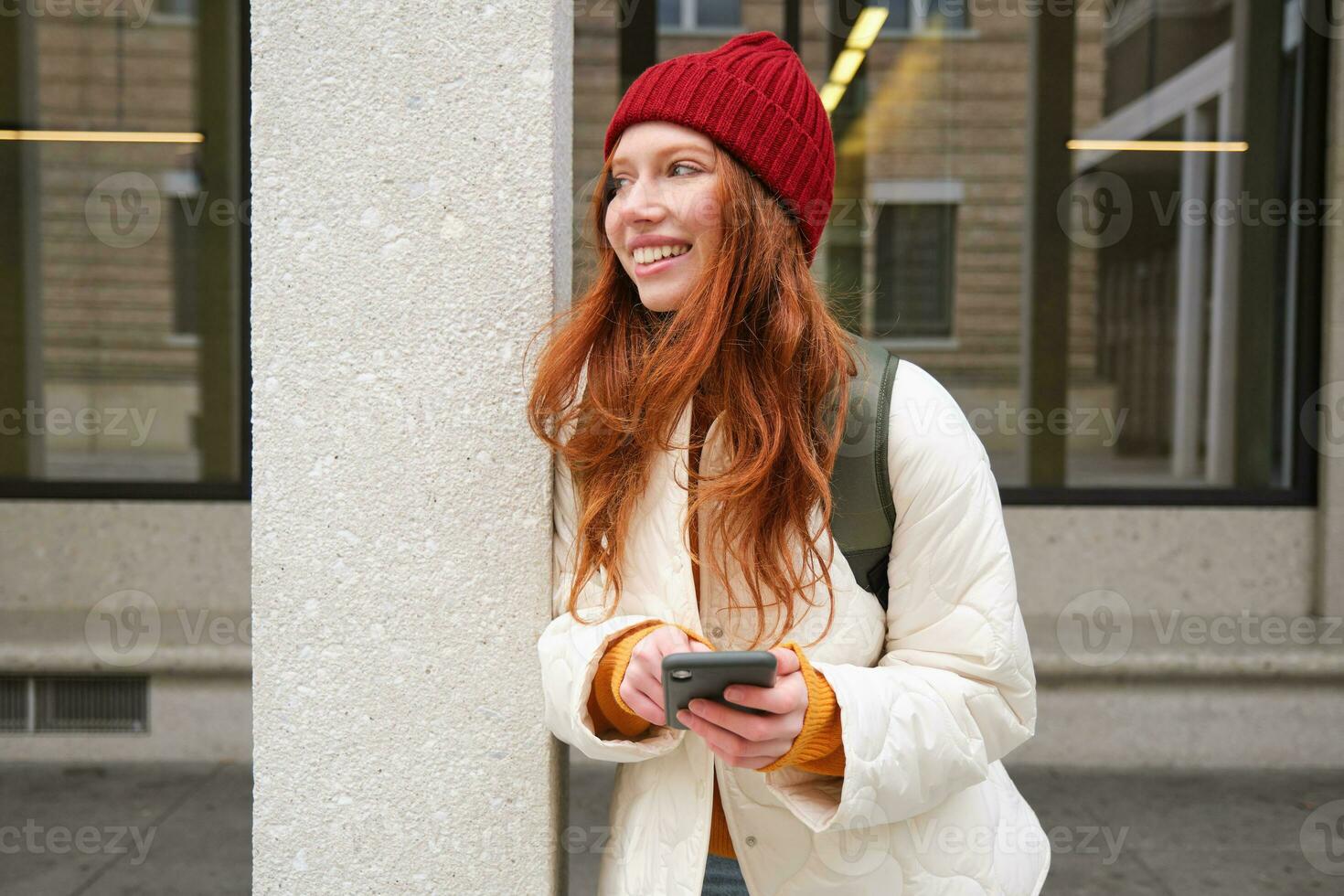 Beautiful redhead girl with smartphone, standing on street, holding mobile phone, using taxi app, connects to public wifi and uses internet, sends a text message photo