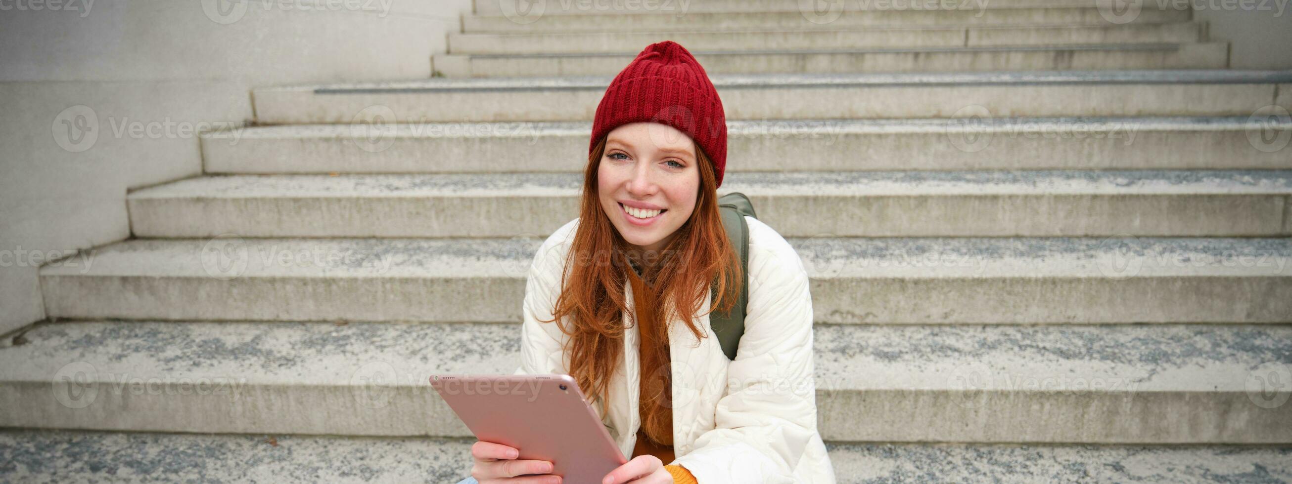 Happy stylish redhead girl, student in red hat, holds digital tablet, uses social media app, searches something online, connects to wifi photo