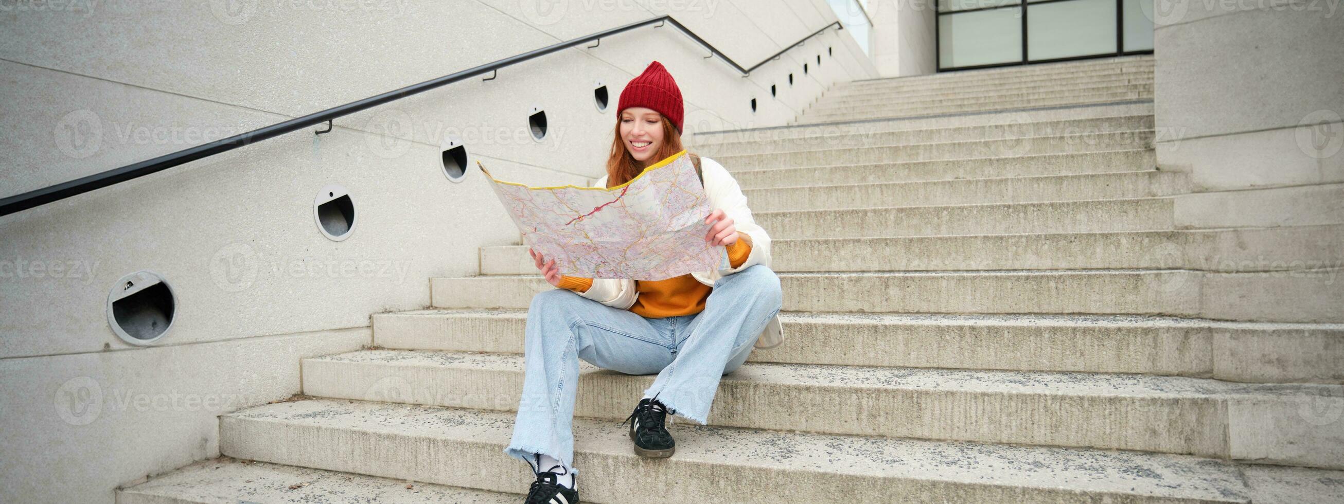 Young smiling redhead girl, tourist sits on stairs outdoors with city paper map, looking for direction, traveller backpacker explores city and looks for sightseeing photo