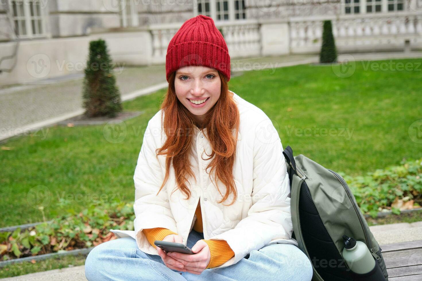joven sonriente pelirrojo niña se sienta en parque con teléfono inteligente, mensajes de texto mensaje en móvil teléfono, utilizando teléfono, conectando a público Wifi y surf Internet foto