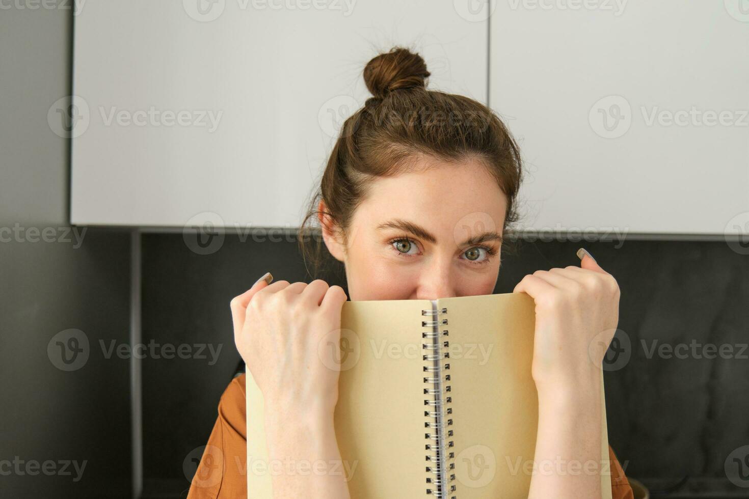 estilo de vida y personas concepto. joven morena mujer, participación cuaderno cerca rostro, ocultación sonrisa detrás bloc y mirando jugado a cámara foto