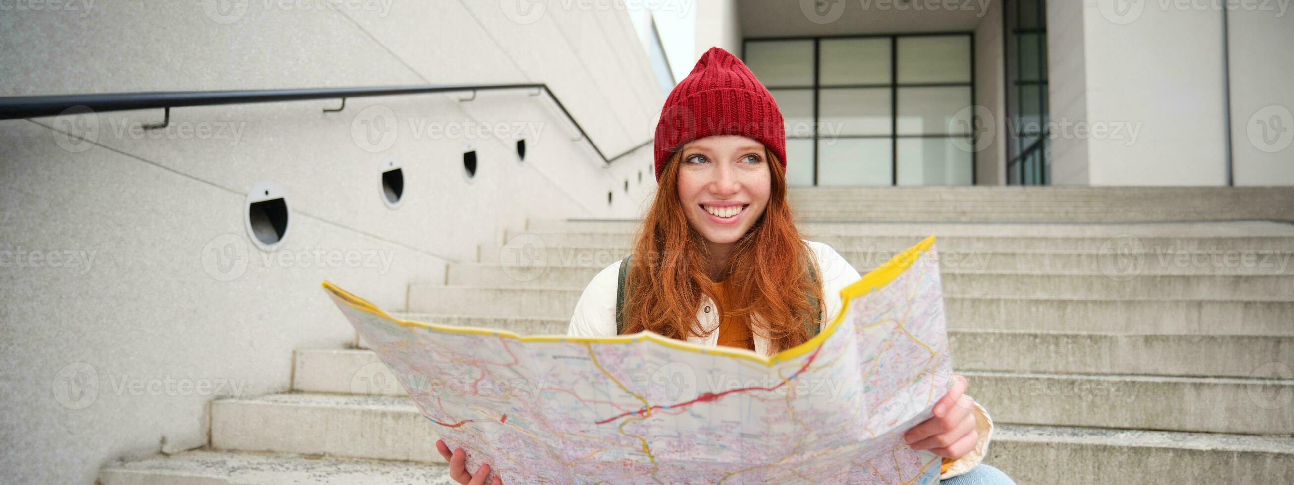 Beautiful girl tourist sits on stairs with city map, plans her journey, looks for direction while travelling around town, searches route for sightseeing photo
