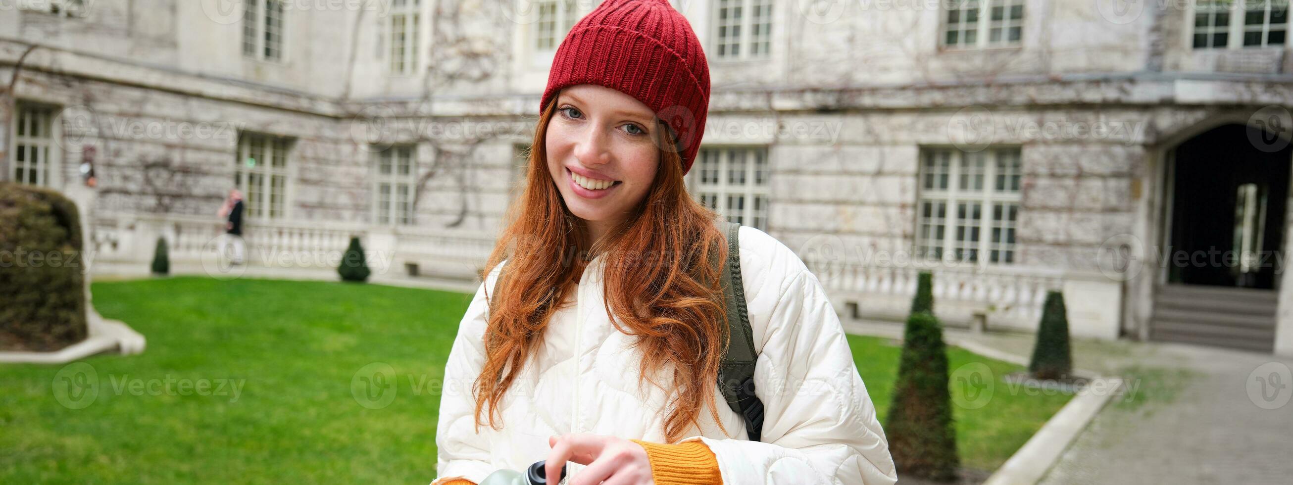 Portrait of happy girl traveler, drinking hot tea from thermos while walking and exploring city. Tourist with flask smiling at camera photo