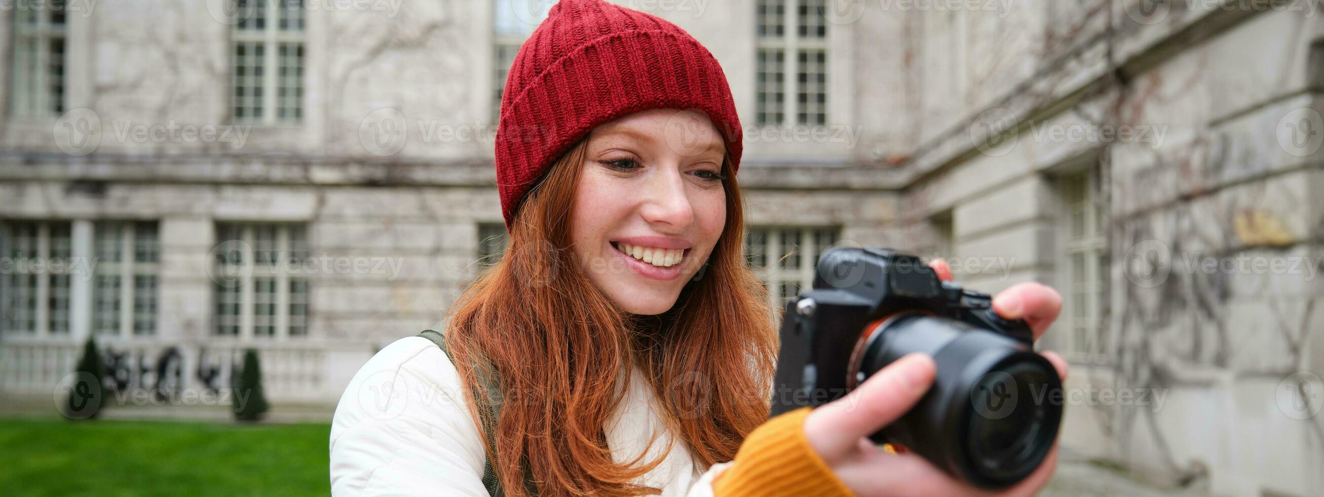 Redhead girl photographer takes photos on professional camera outdoors, captures streetstyle shots, looks excited while taking pictures