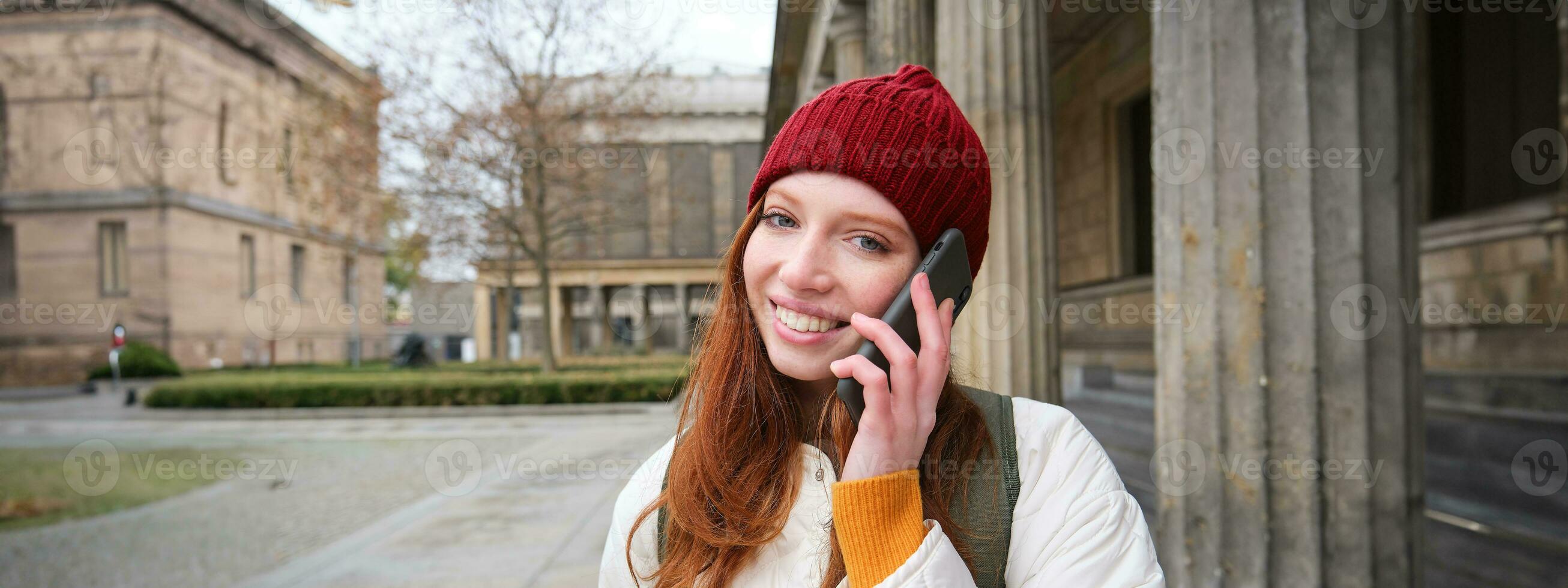 sonriente joven pelirrojo mujer escucha a voz mensaje, hace un teléfono llamar, camina en calle y negociaciones a alguien en teléfono inteligente foto