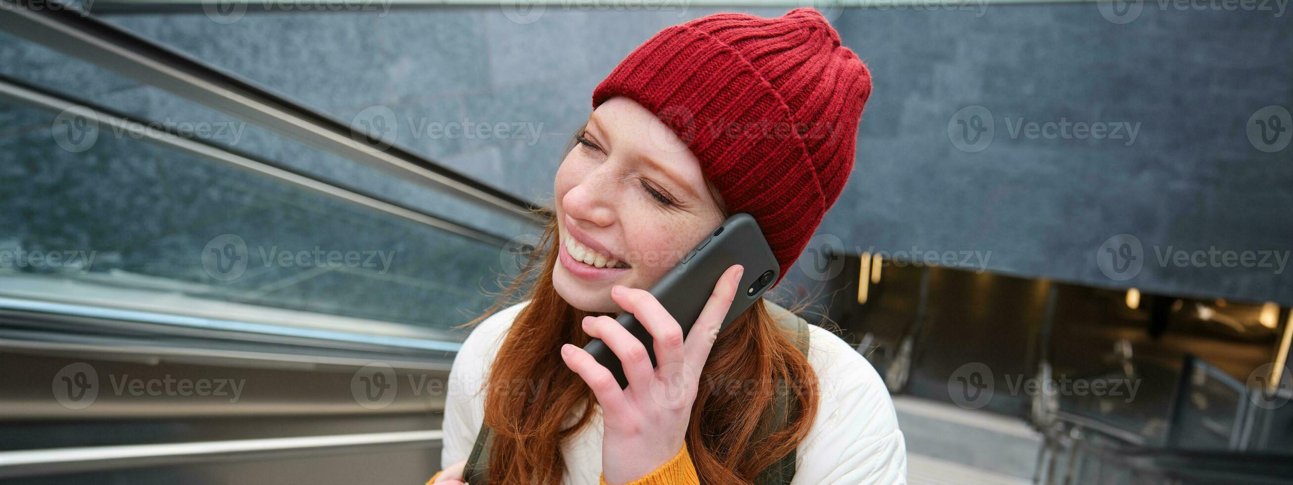 retrato de contento pelirrojo mujer caminando alrededor pueblo con teléfono inteligente, vocación alguien, hablando en móvil teléfono al aire libre foto