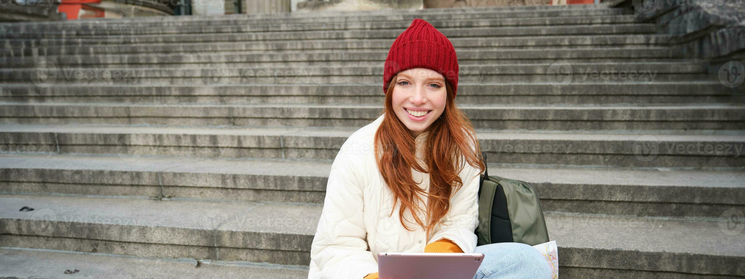 Beautiful young modern girl with red hair, holds digital tablet, sits on stairs near museum and connects public internet, sends message on gadget app photo