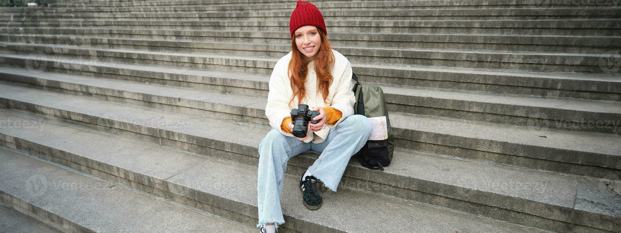 Young student, photographer sits on street stairs and checks her shots on professional camera, taking photos outdoors