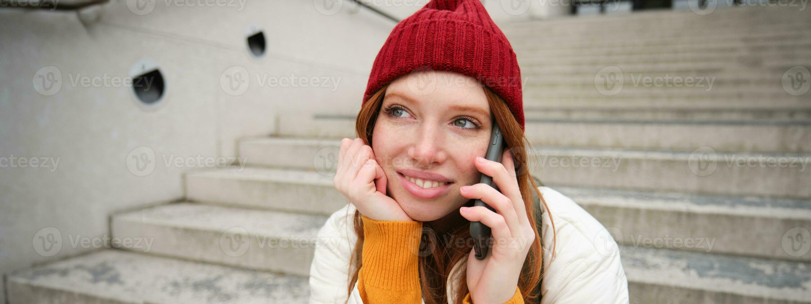 hermosa sonriente pelirrojo hembra modelo, se sienta en calle y negociaciones en móvil teléfono, usos teléfono inteligente aplicación a llamada en el extranjero, riendo durante teléfono conversacion foto