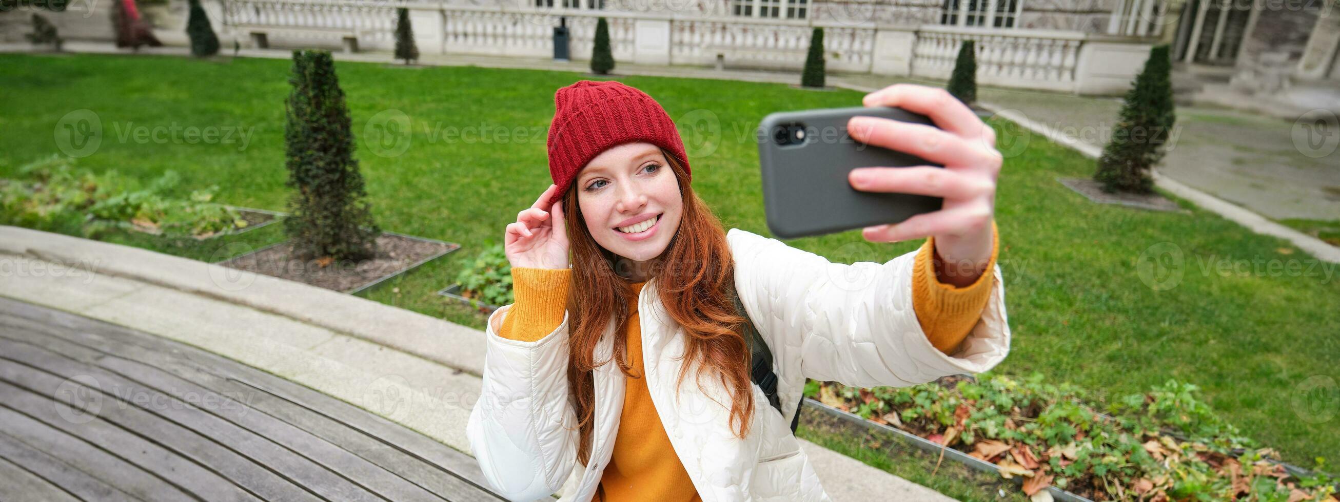joven adolescente pelirrojo niña se sienta en banco en parque y toma autofoto, hace un foto de sí misma con teléfono inteligente aplicación, registros vlog