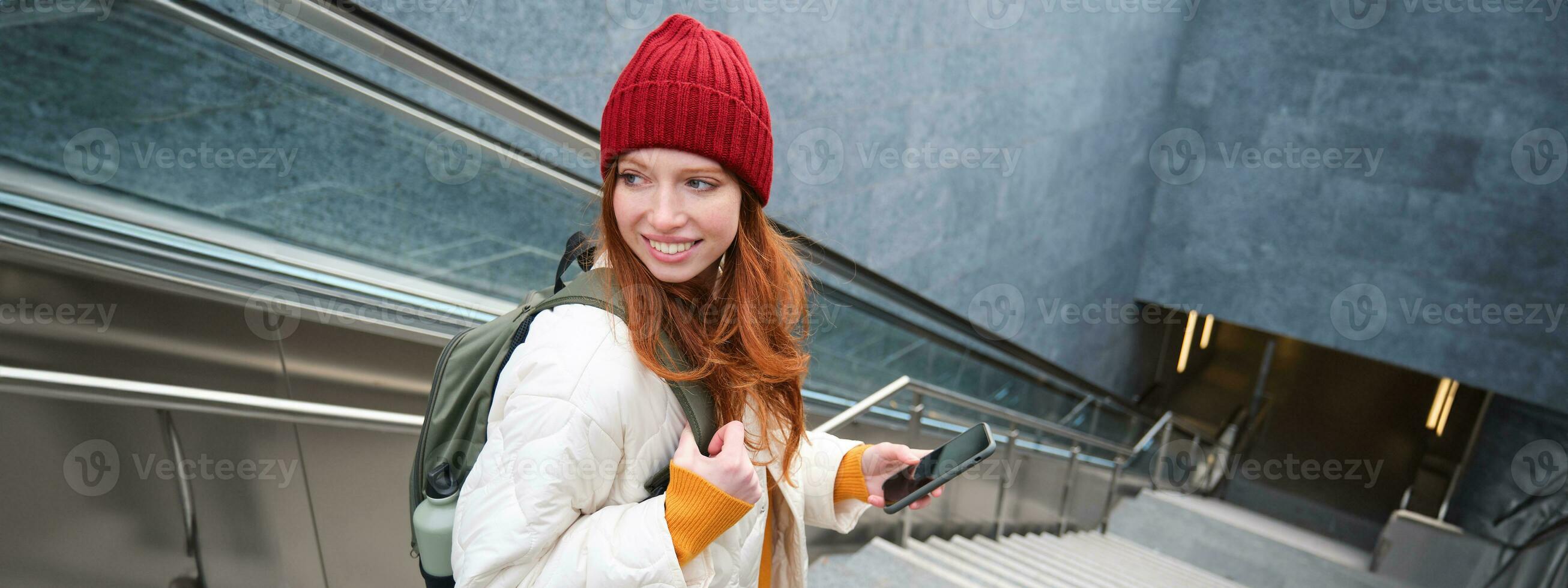 Portrait of redhead girl exploring city with smartphone map app. Young woman using mobile phone and walking around city, going down stairs photo