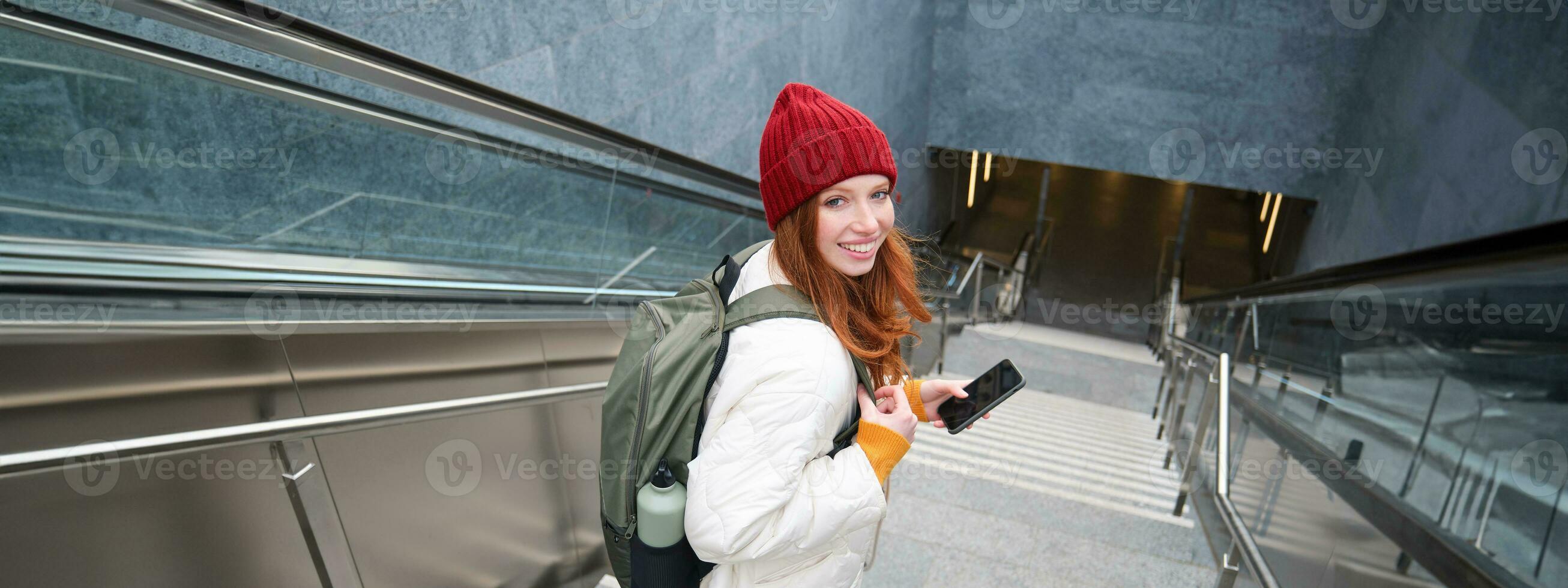 Portrait of redhead girl exploring city with smartphone map app. Young woman using mobile phone and walking around city, going down stairs photo