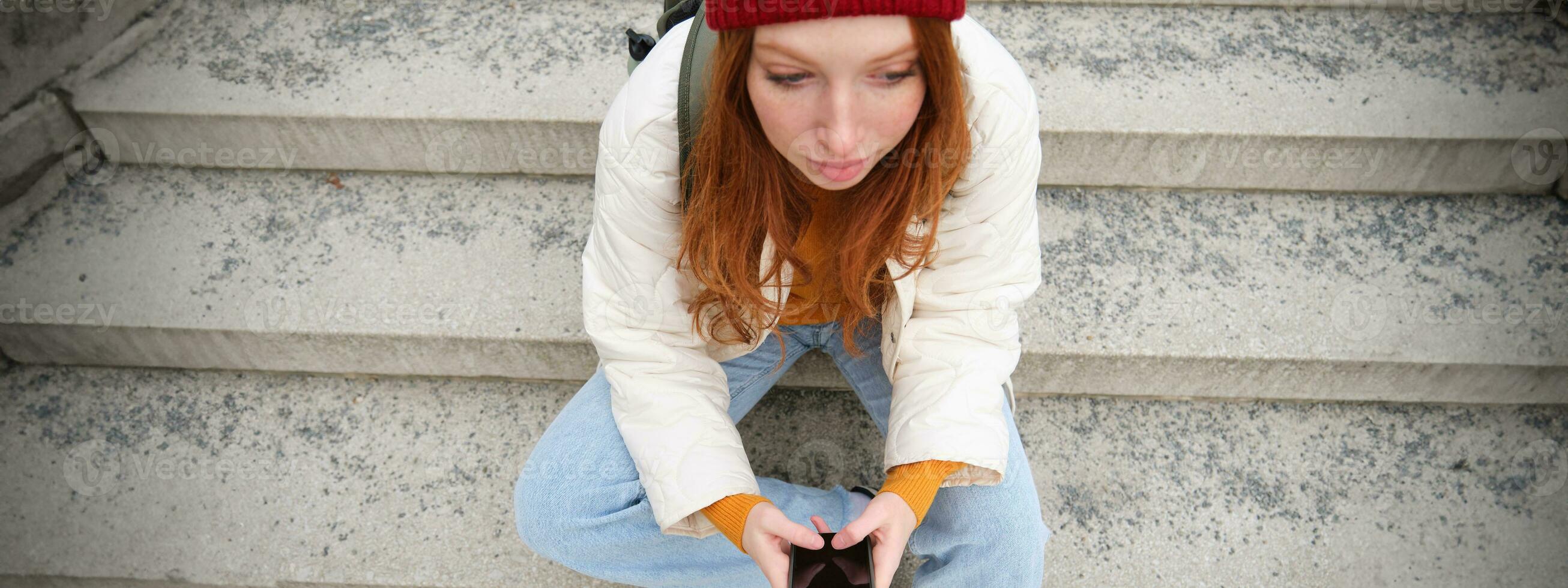 High angle photo of stylish redhead teen girl texts message on phone, uses mobile application while sits outdoors on public stairs