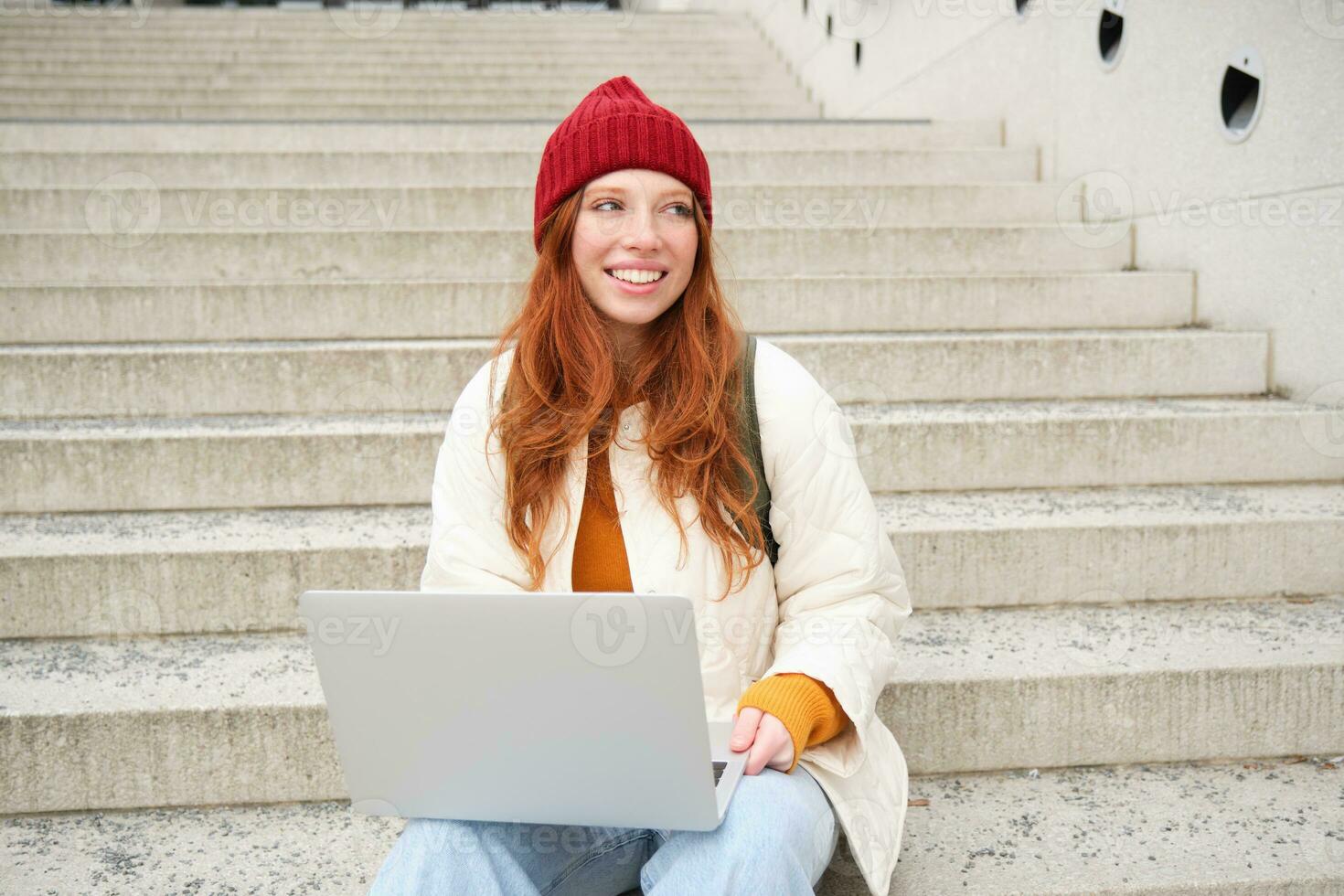Stylish young redhead girl, student with laptop sitting on stairs near building, connects to public wifi and works on project, chats on computer photo