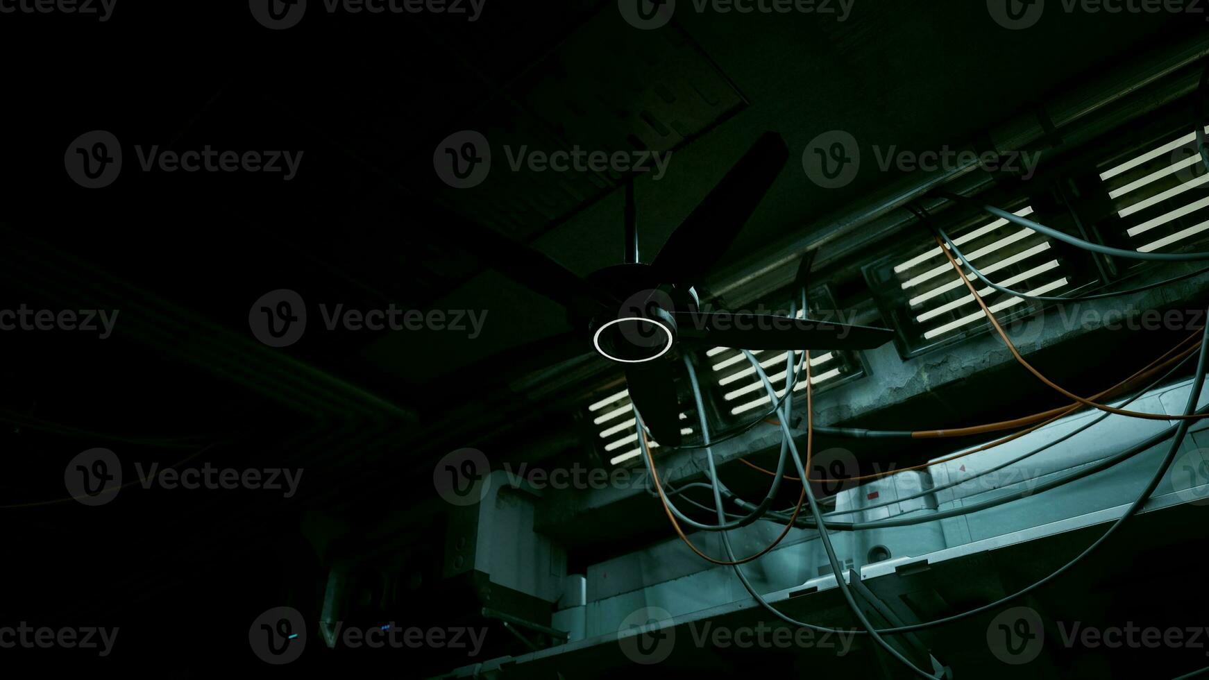 A dark concrete interior with a ceiling fan and multiple wires hanging from it photo