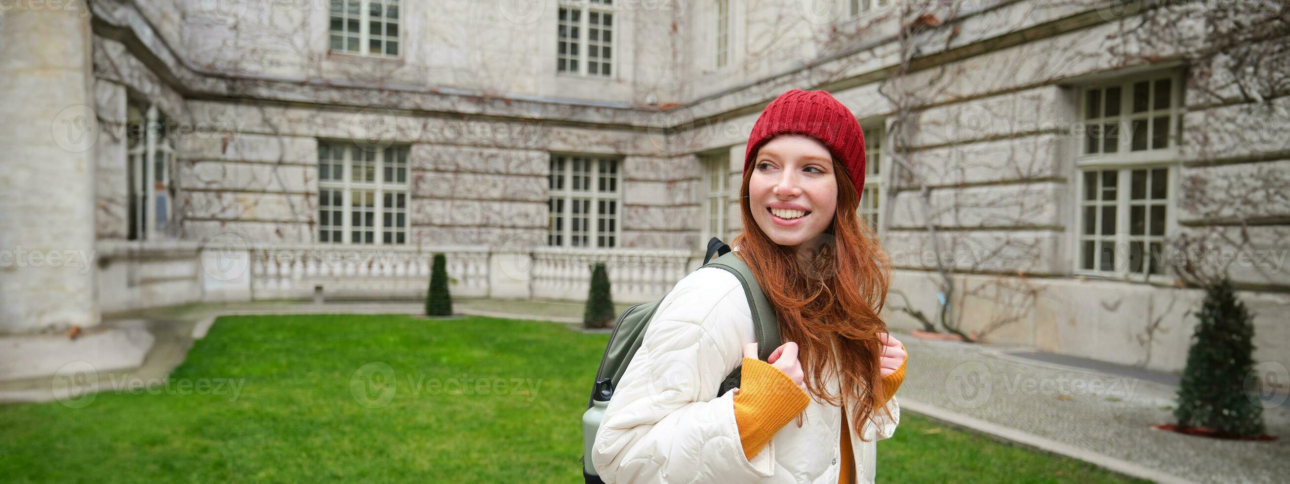 Beautiful redhead woman with backpack, tourist walking around city park and looks around, wears red hat and warm coat photo