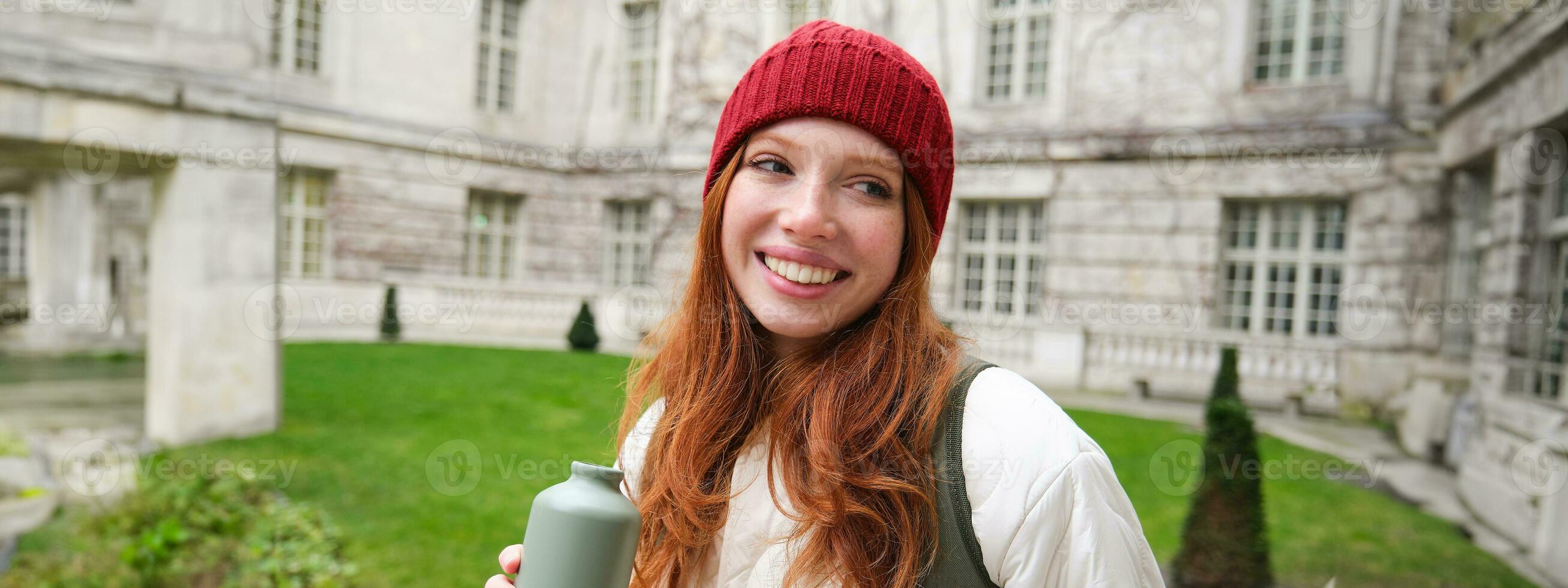 retrato de contento joven mujer, turista con mochila pasear, Bebiendo caliente té desde termo, participación matraz y sonriente foto