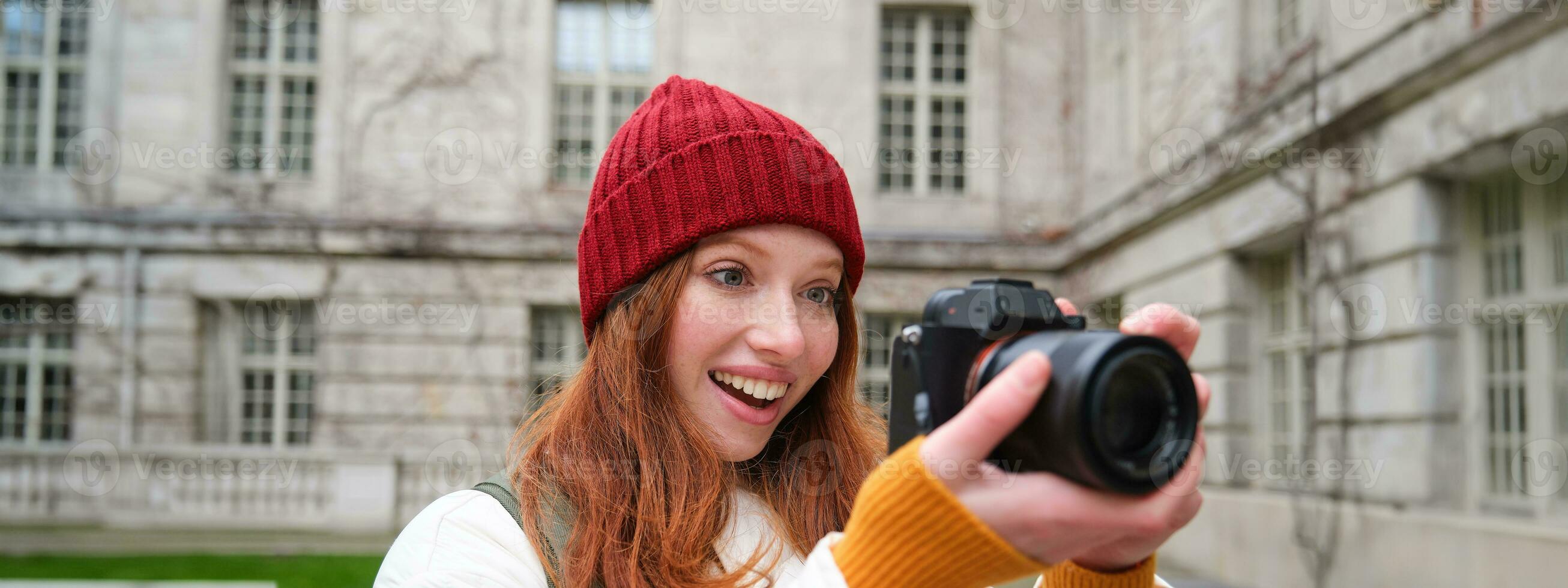 Redhead girl photographer takes photos on professional camera outdoors, captures streetstyle shots, looks excited while taking pictures