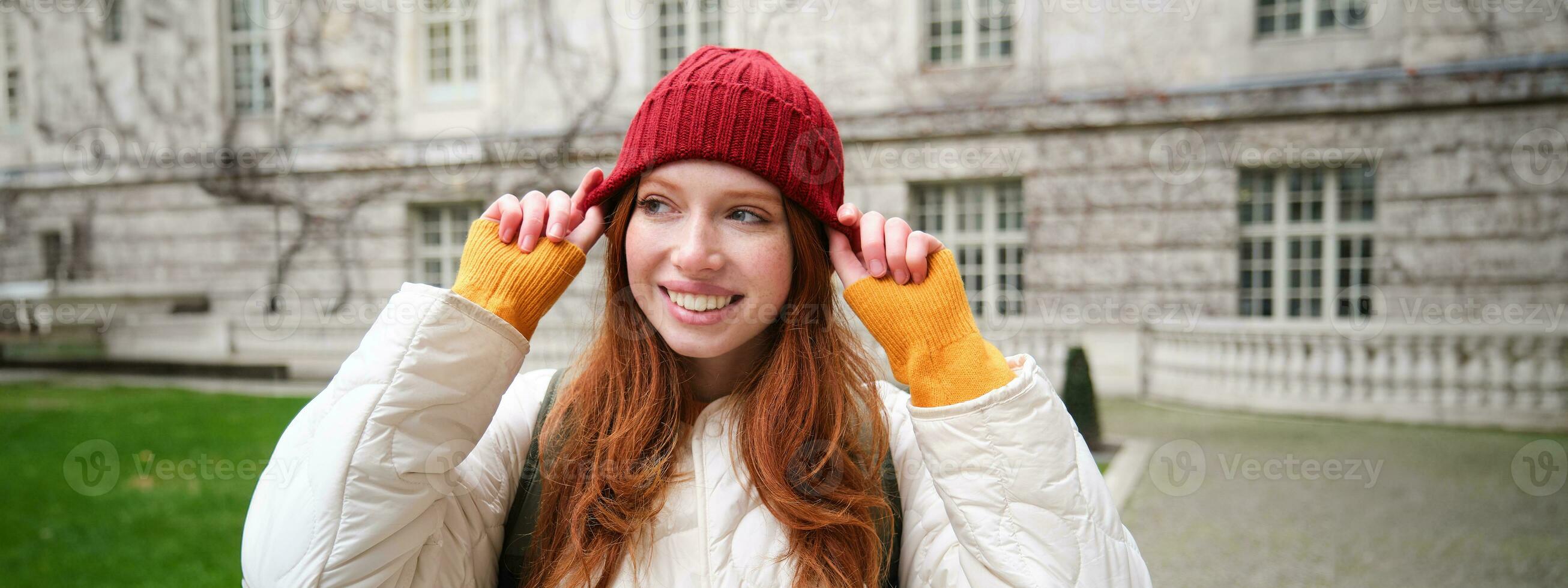 Portrait of smiling redhead woman puts on red hat and smiles, wearing warm clothes while exploring city streets photo