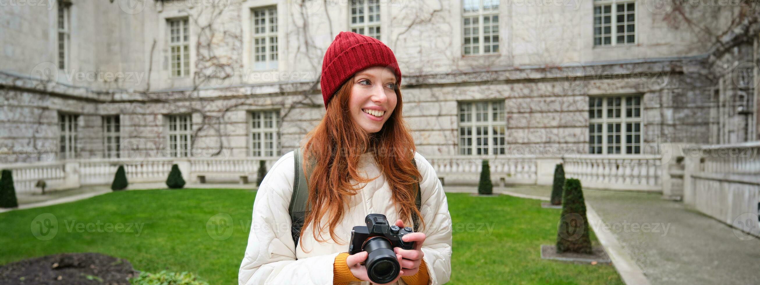 Redhead girl photographer takes photos on professional camera outdoors, captures streetstyle shots, looks excited while taking pictures