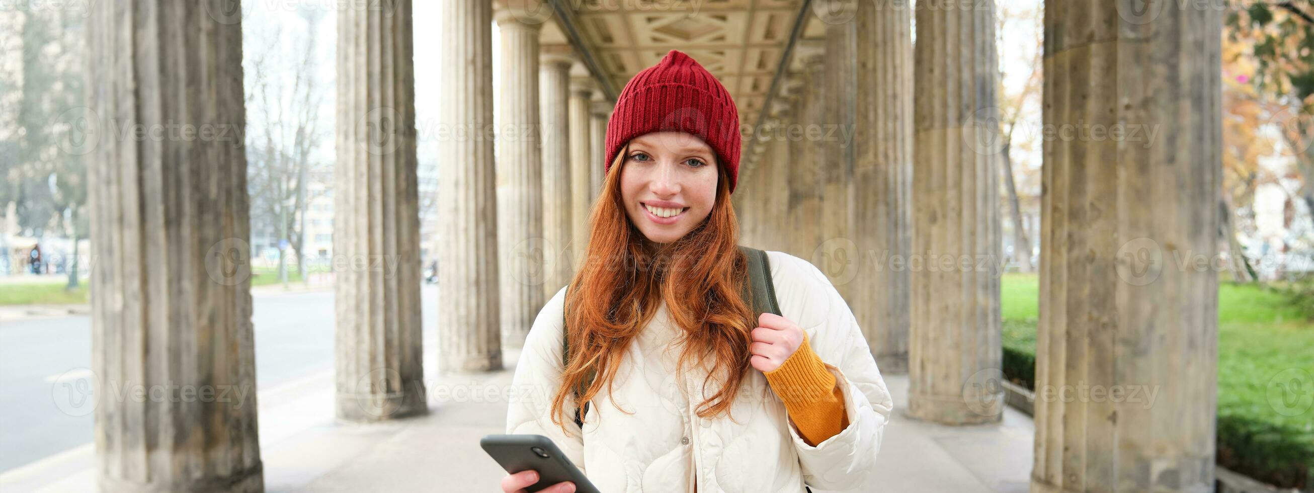 Happy young female model stands on street and holds mobile phone, uses smartphone app, follows the route of online map photo