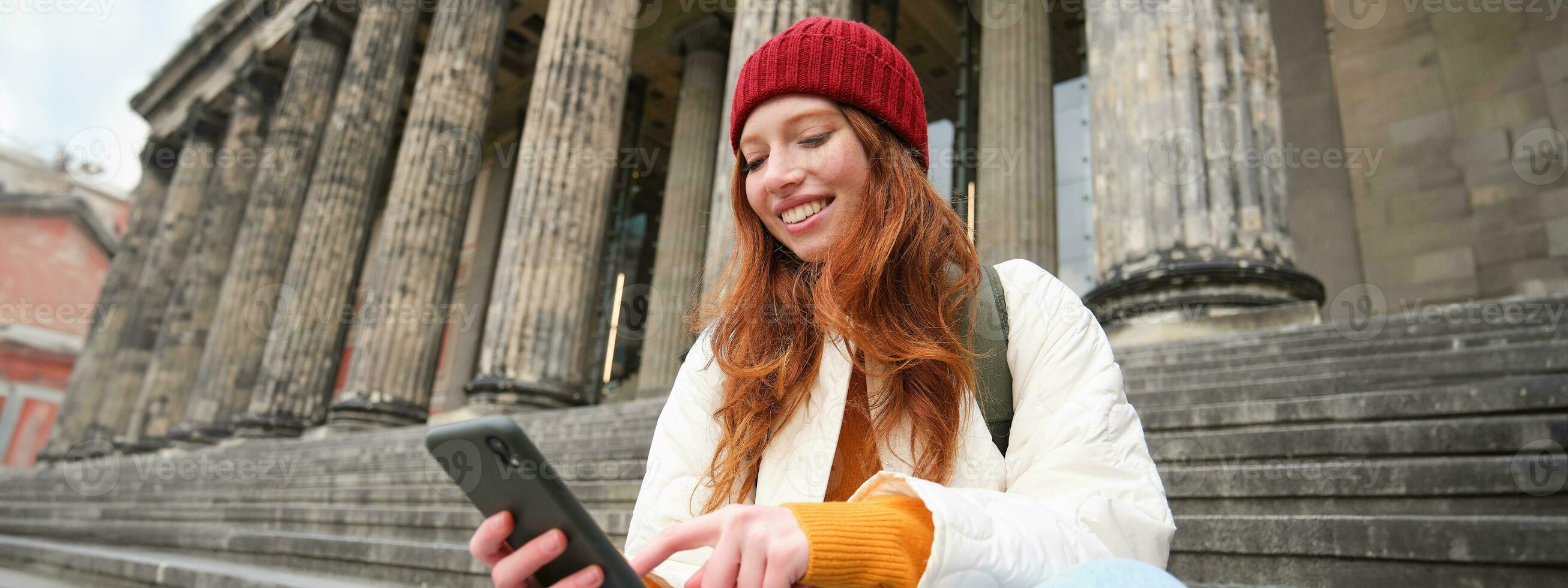 Stylish young redhead woman, talking on mobile phone app, using social media application, looking for something online on smartphone, sits on stairs outdoors photo
