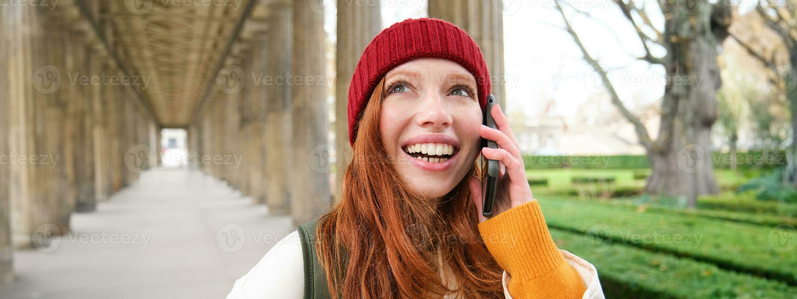 retrato de pelirrojo europeo niña en rojo sombrero, hace un teléfono llamar, camina en ciudad y negociaciones a amigo en teléfono inteligente foto