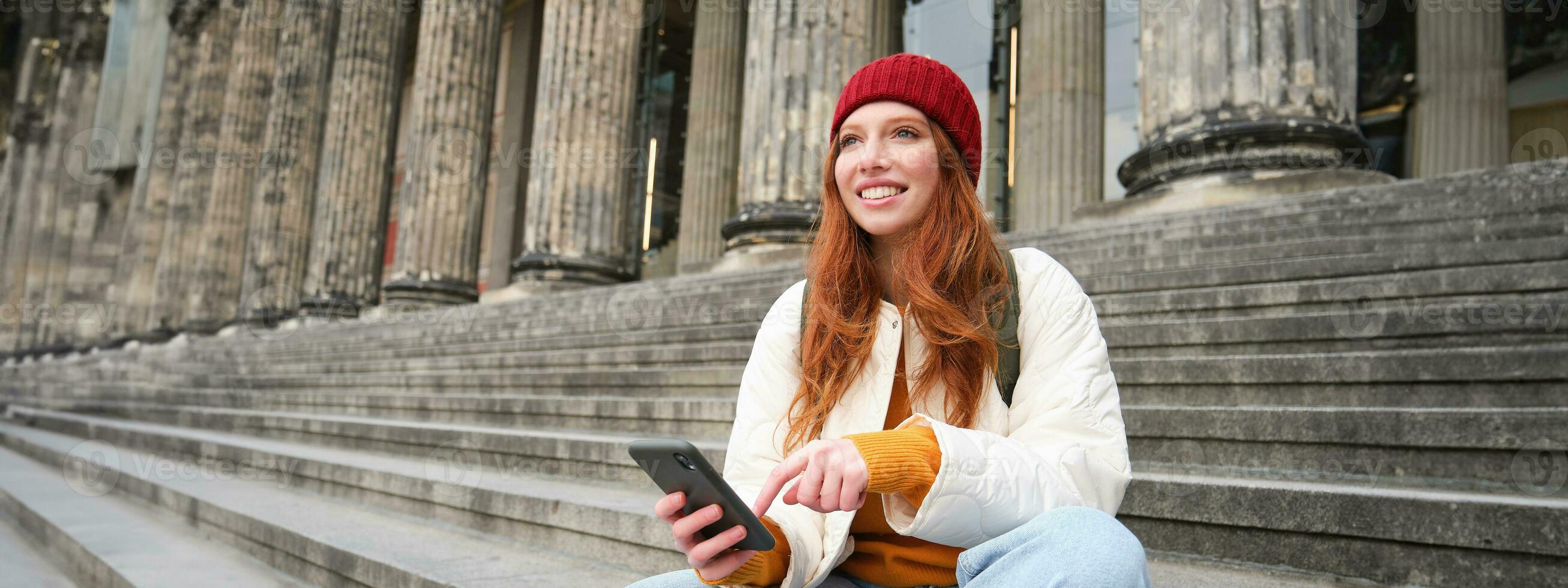 Portrait of young urban girl in red hat, sits on stairs near museum, holds mobile phone, connects to public wifi and surfs net, uses smartphone apps photo