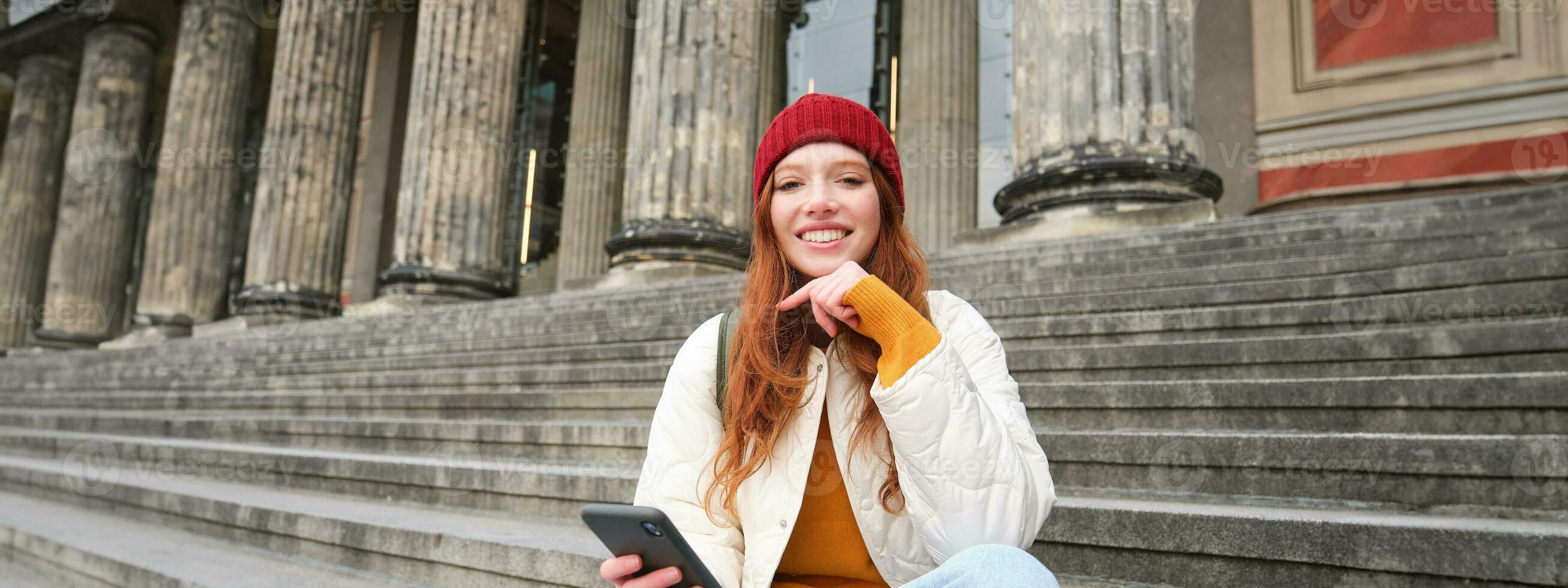 Portrait of young urban girl in red hat, sits on stairs near museum, holds mobile phone, connects to public wifi and surfs net, uses smartphone apps photo