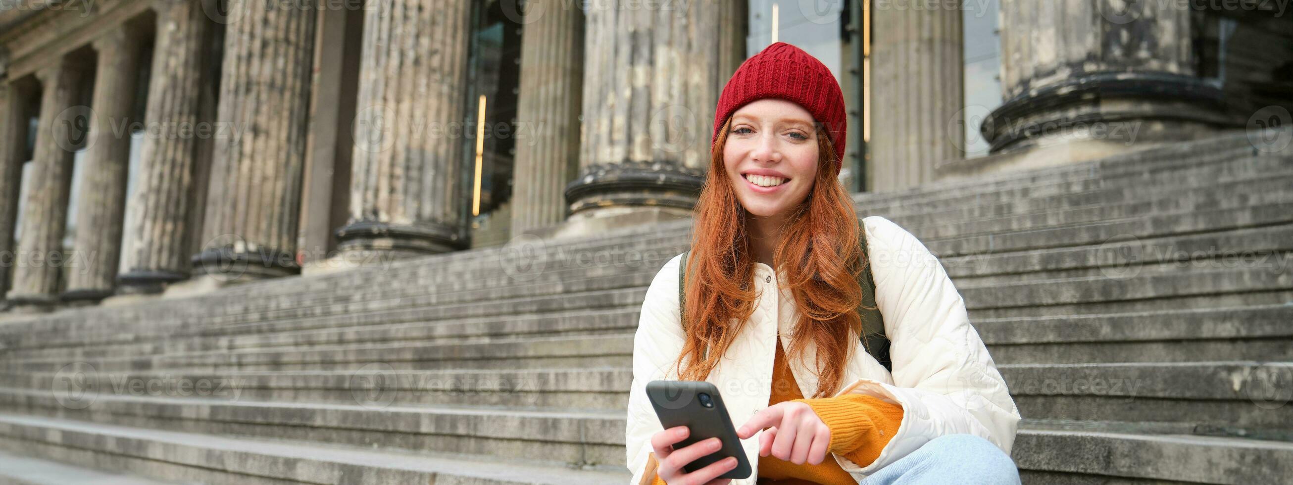 Portrait of young urban girl in red hat, sits on stairs near museum, holds mobile phone, connects to public wifi and surfs net, uses smartphone apps photo