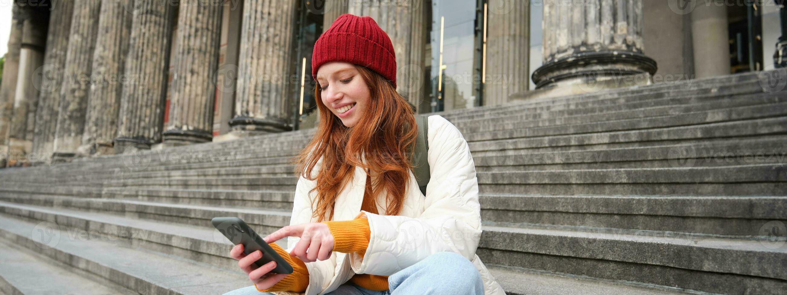 Portrait of young urban girl in red hat, sits on stairs near museum, holds mobile phone, connects to public wifi and surfs net, uses smartphone apps photo