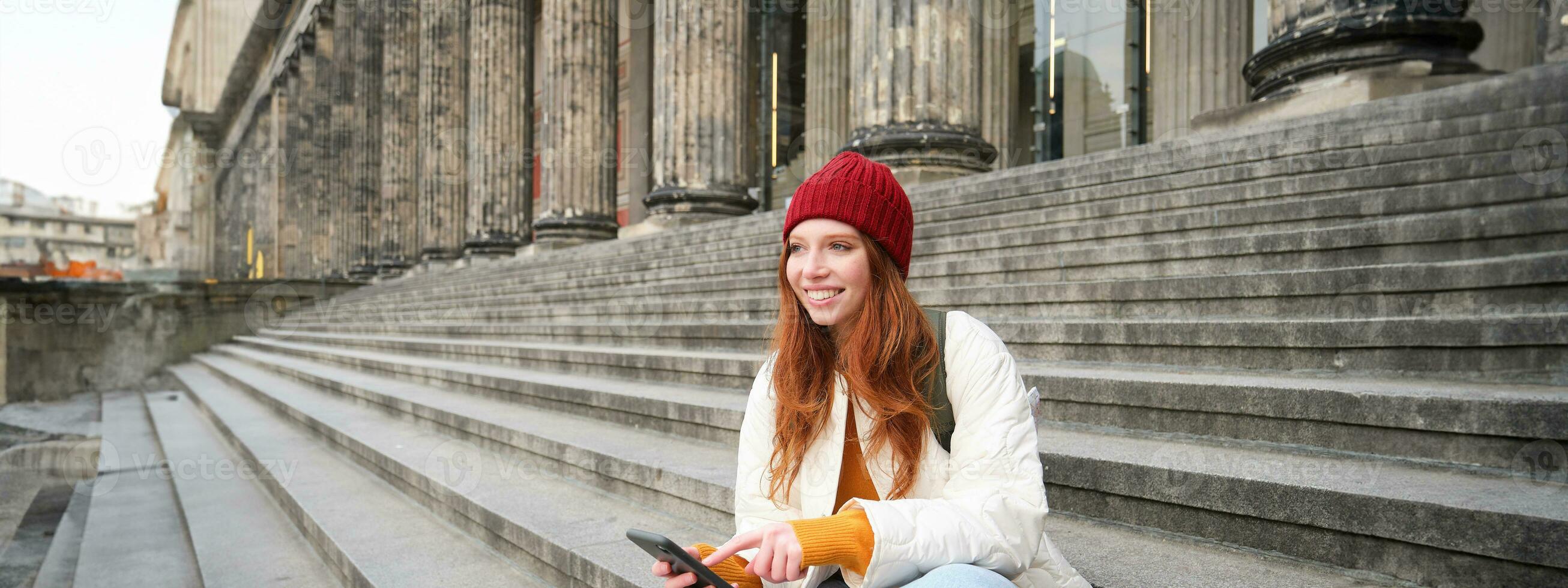 Stylish young redhead woman, talking on mobile phone app, using social media application, looking for something online on smartphone, sits on stairs outdoors photo