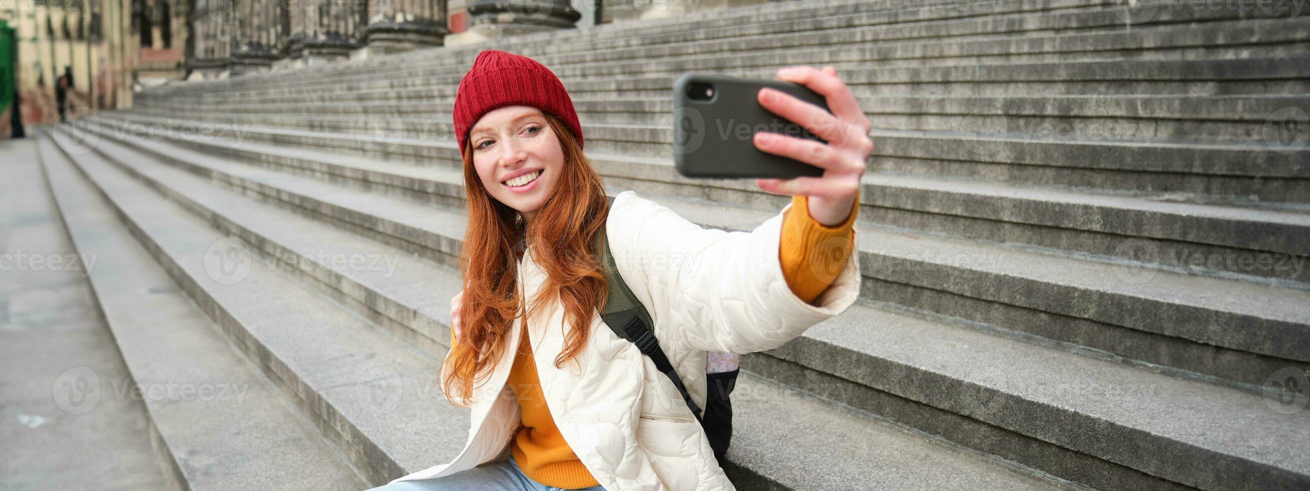 joven pelirrojo turista toma selfie en frente de museo en escaleras, sostiene teléfono inteligente y mira a móvil cámara, hace foto de sí misma con teléfono