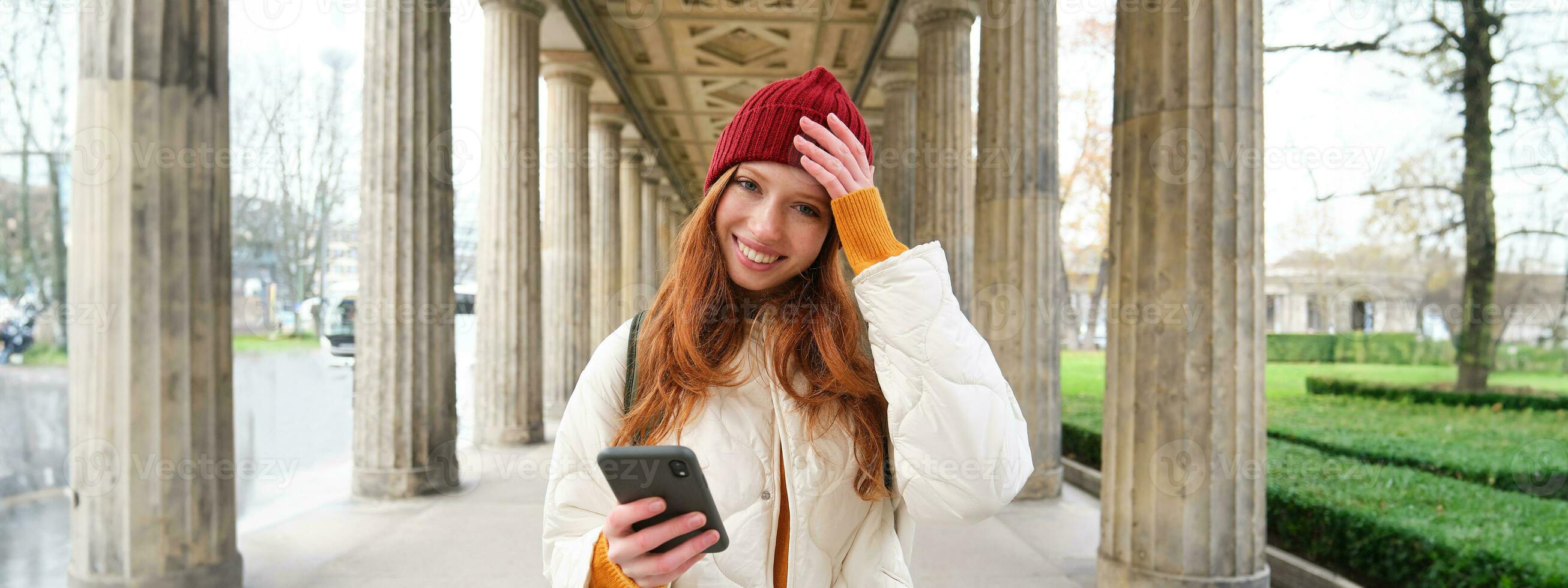 Happy young female model stands on street and holds mobile phone, uses smartphone app, follows the route of online map photo
