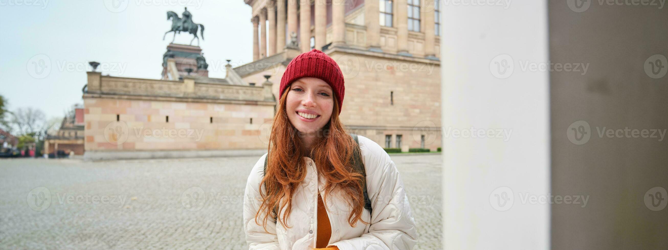 Smiling redhead girl tourist, walks around city and explores popular landmarks, sightseeing, holding smartphone, looking at her mobile app and checks with city map photo