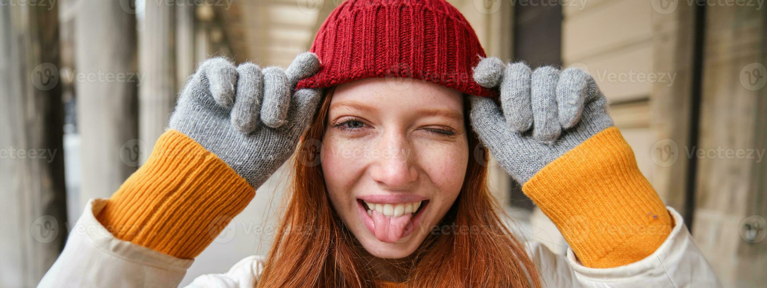 retrato de joven pelirrojo mujer en de punto sombrero y guantes, sonrisas y mira aparte, camina alrededor ciudad en invierno foto