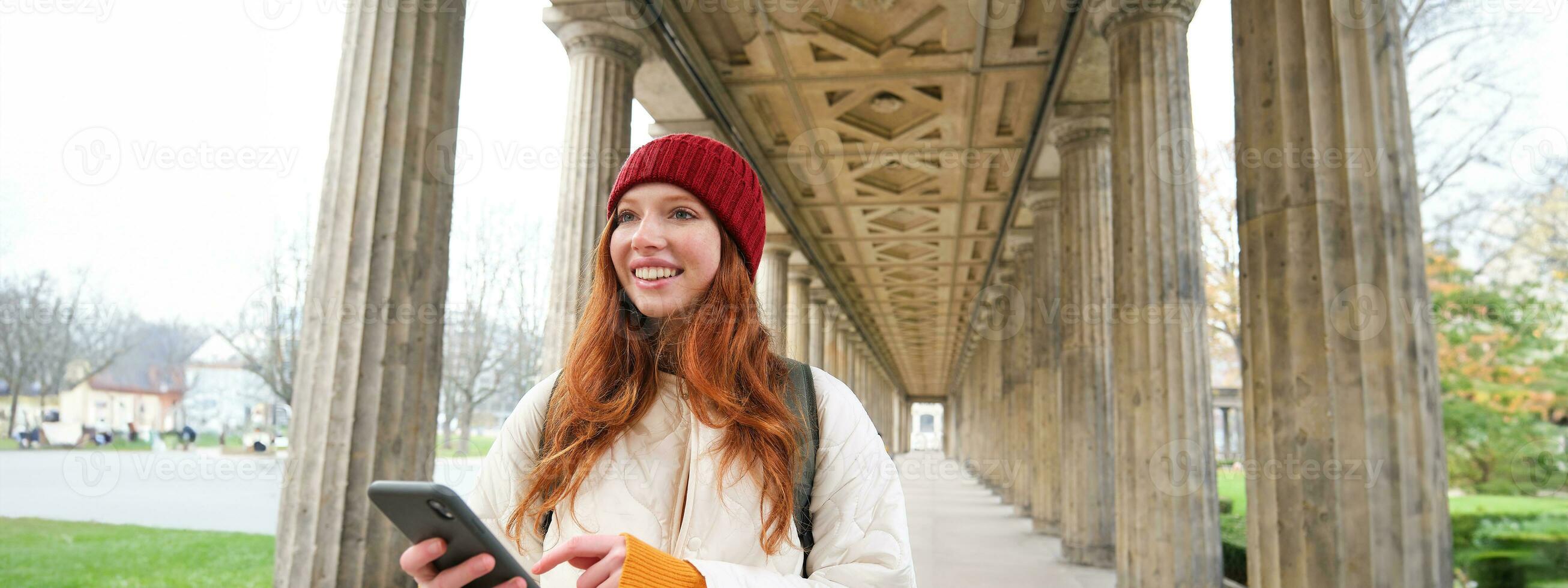 Happy young female model stands on street and holds mobile phone, uses smartphone app, follows the route of online map photo