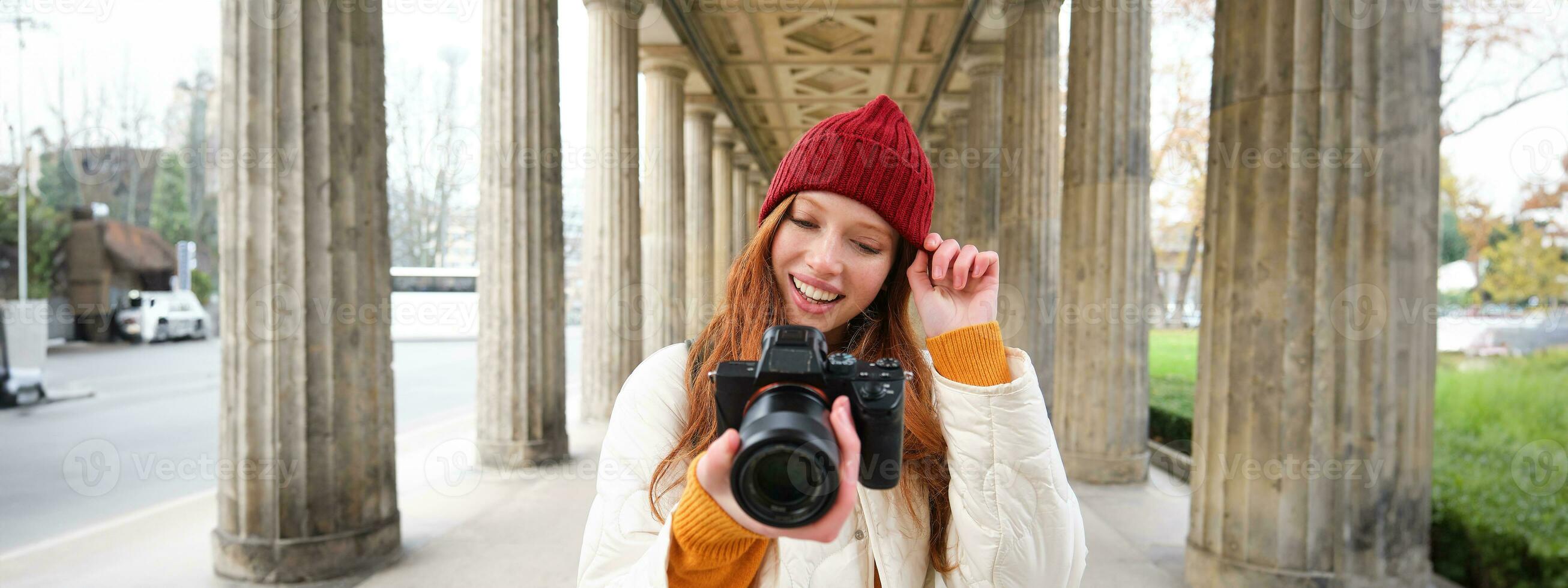 joven pelirrojo hembra fotógrafo, hace estilo de vida disparo en ciudad centro, toma fotos y sonrisas, mira para Perfecto disparo, hace imagen