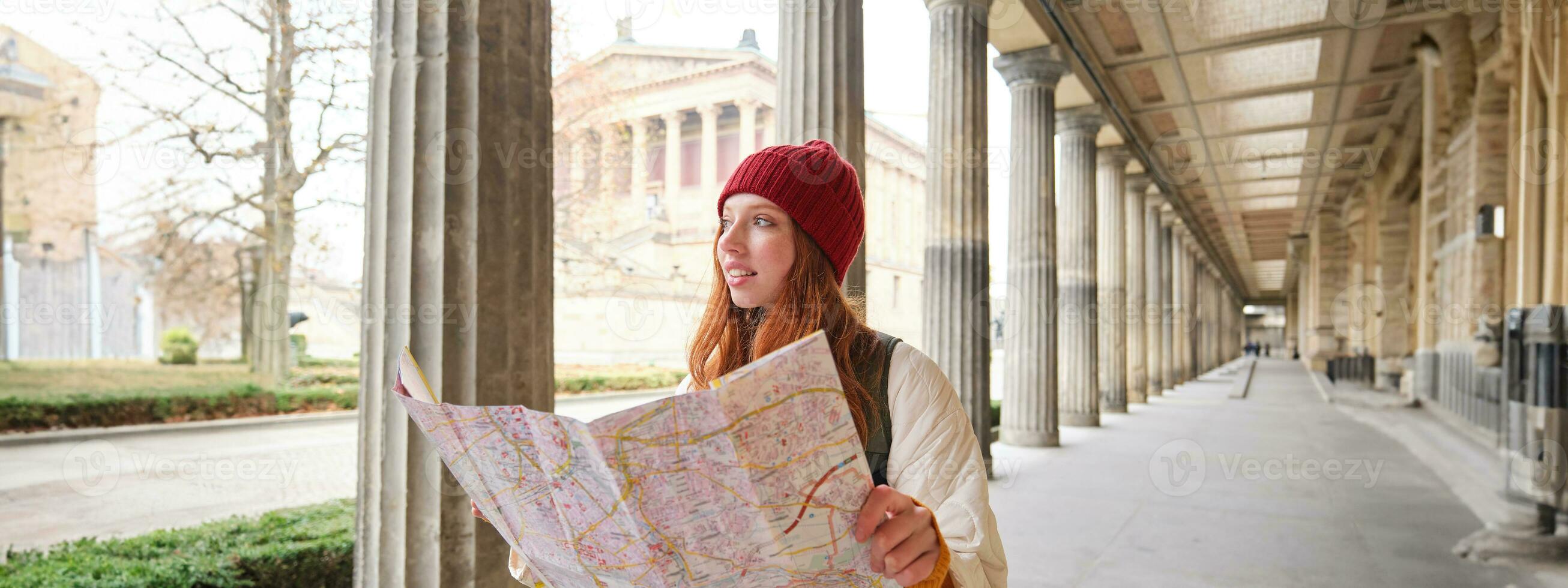 Smiling young redhead woman in red hat, looks at paper map to look for tourist attraction. Tourism and people concept. Girl explores city, tried to find way photo