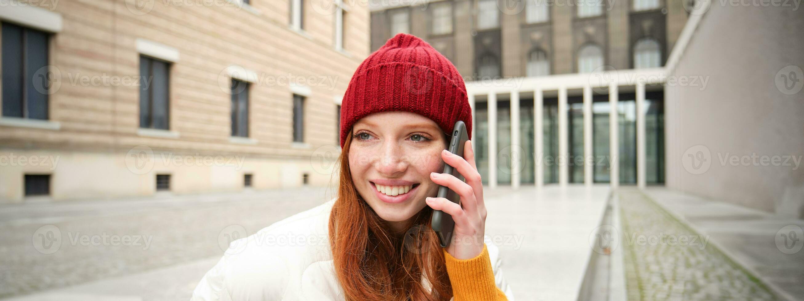 elegante moderno pelirrojo niña negociaciones en móvil teléfono, hace un teléfono llamar, vocación alguien en teléfono inteligente aplicación desde afuera, soportes en calle y sonrisas foto