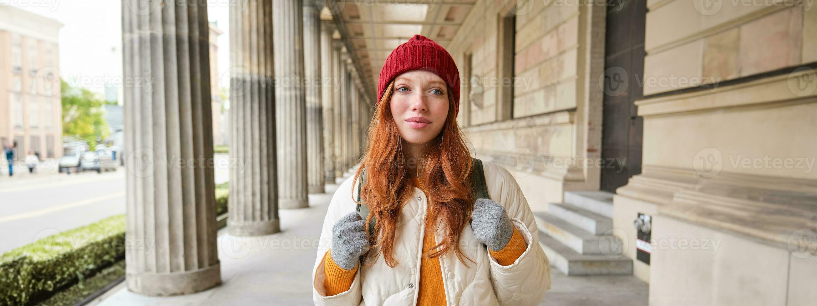 Smiling redhead girl with backpack, walks in city and does sightseeing, explore popular landmarks on her tourist journey around Europe photo