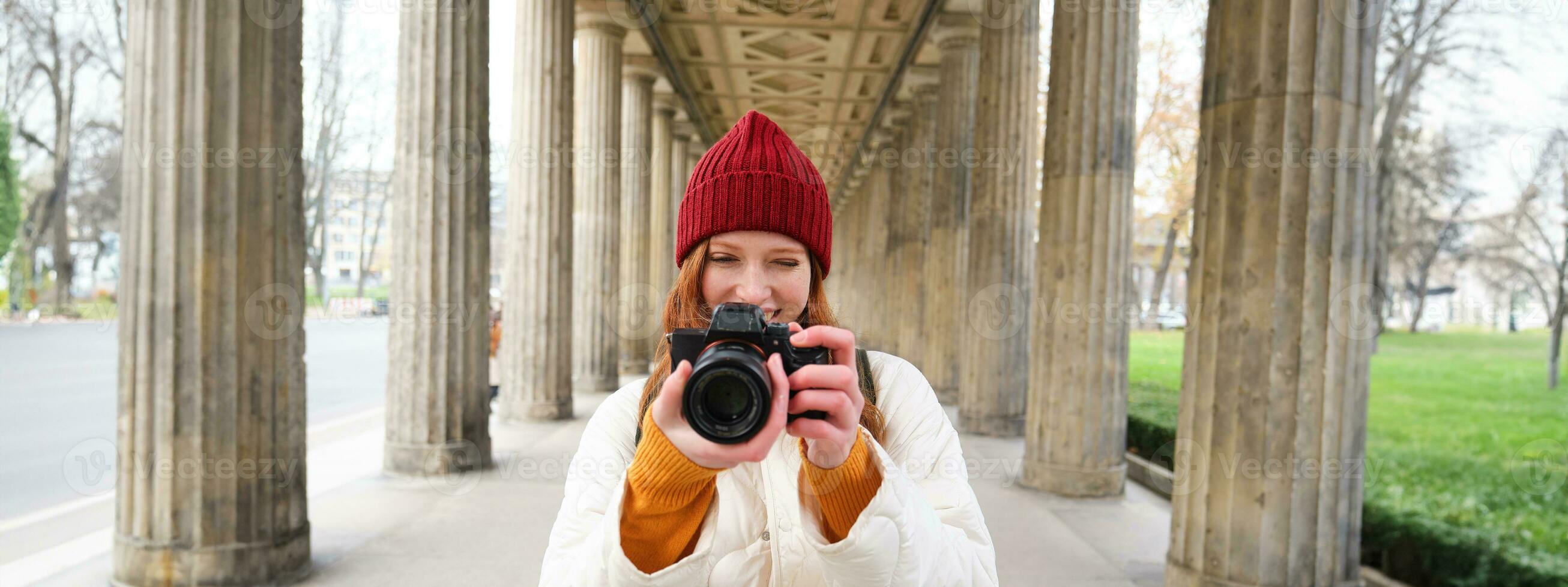 Young redhead female photographer, makes lifestyle shooting in city centre, takes photos and smiles, looks for perfect shot, makes picture