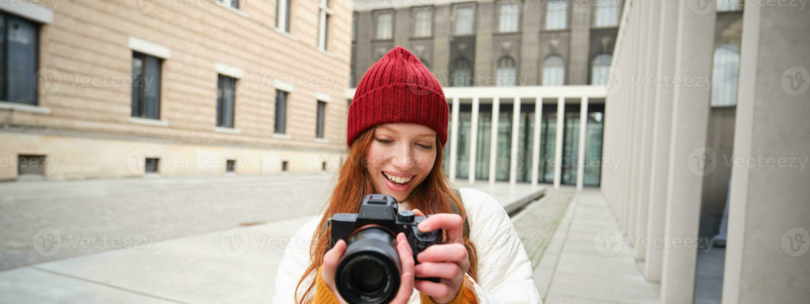 Smiling redhead girl photographer, taking pictures in city, makes photos outdoors on professional camera