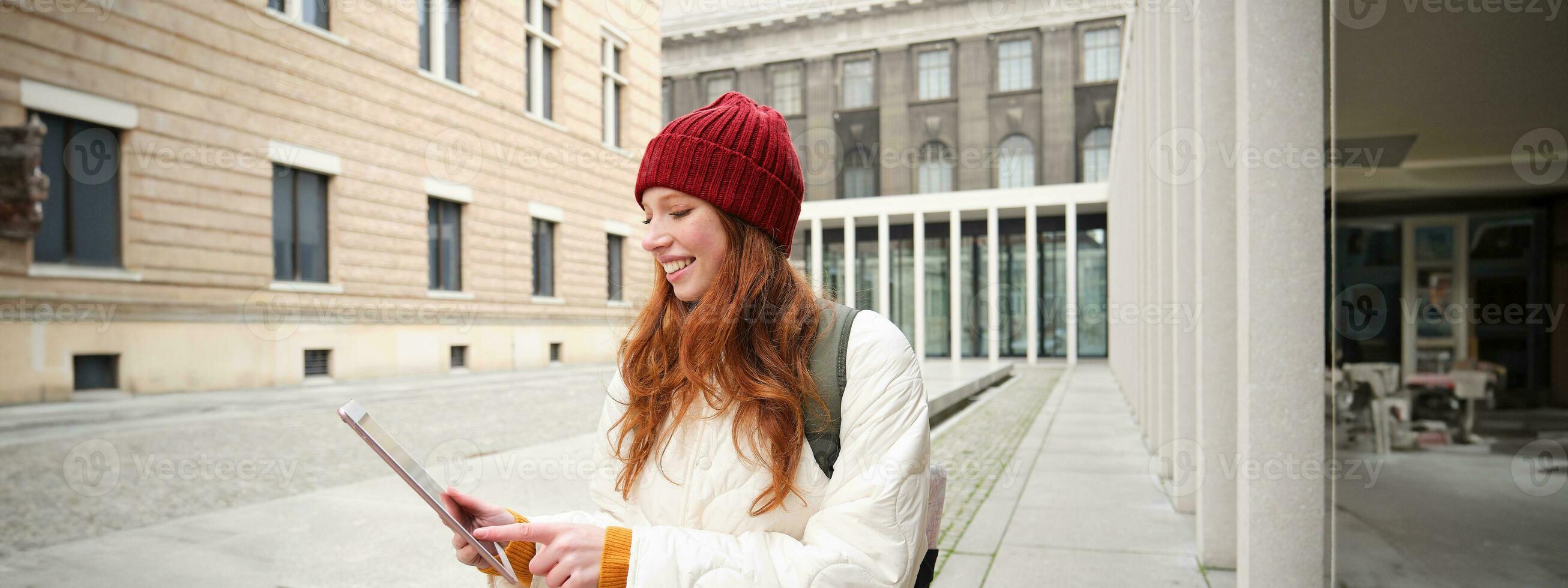 Happy redhead girl in red hat, walks around city with digital tablet, connects to public internet wifi and looks for route, looks at map on her gadget photo