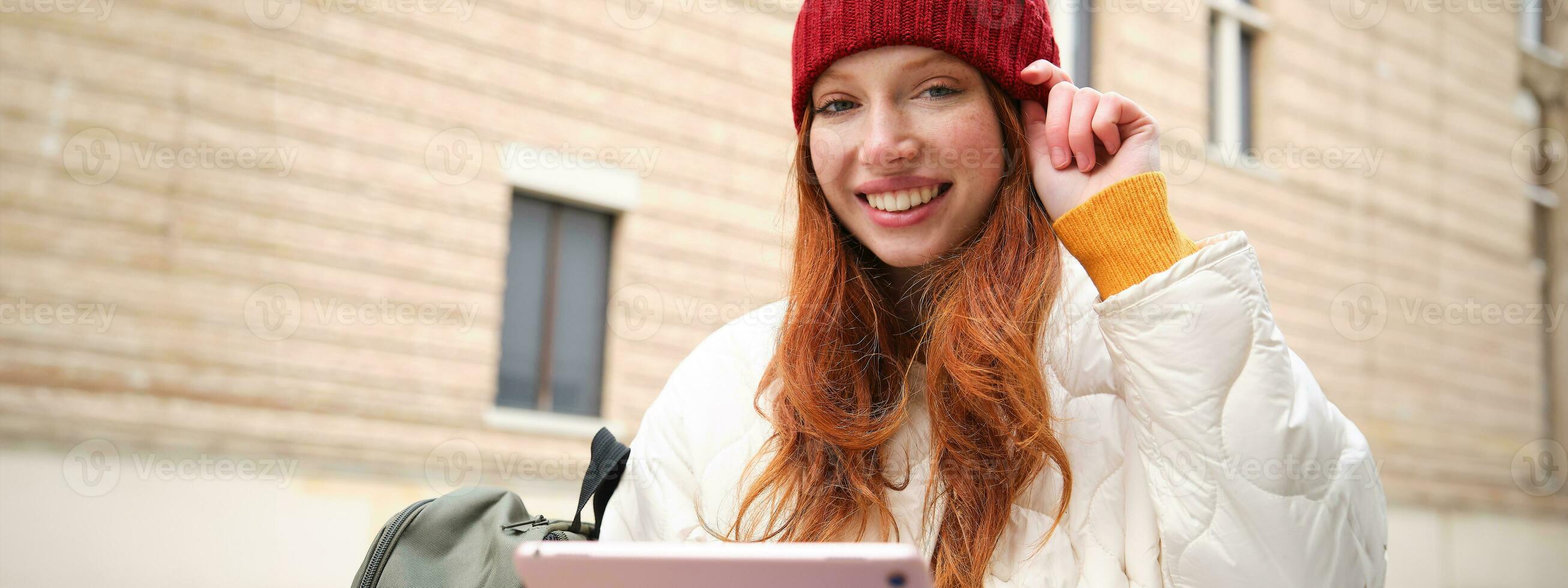 Beautiful redhead woman in red hat, sits with backpack and thermos, using digital tablet outdoors, connects to wifi, texts message, books tickets online photo