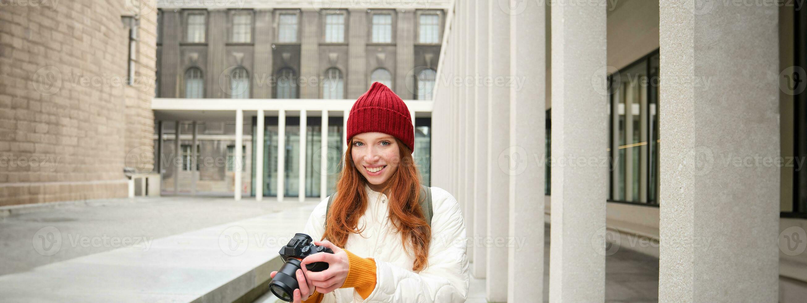 Smiling redhead girl photographer, taking pictures in city, makes photos outdoors on professional camera