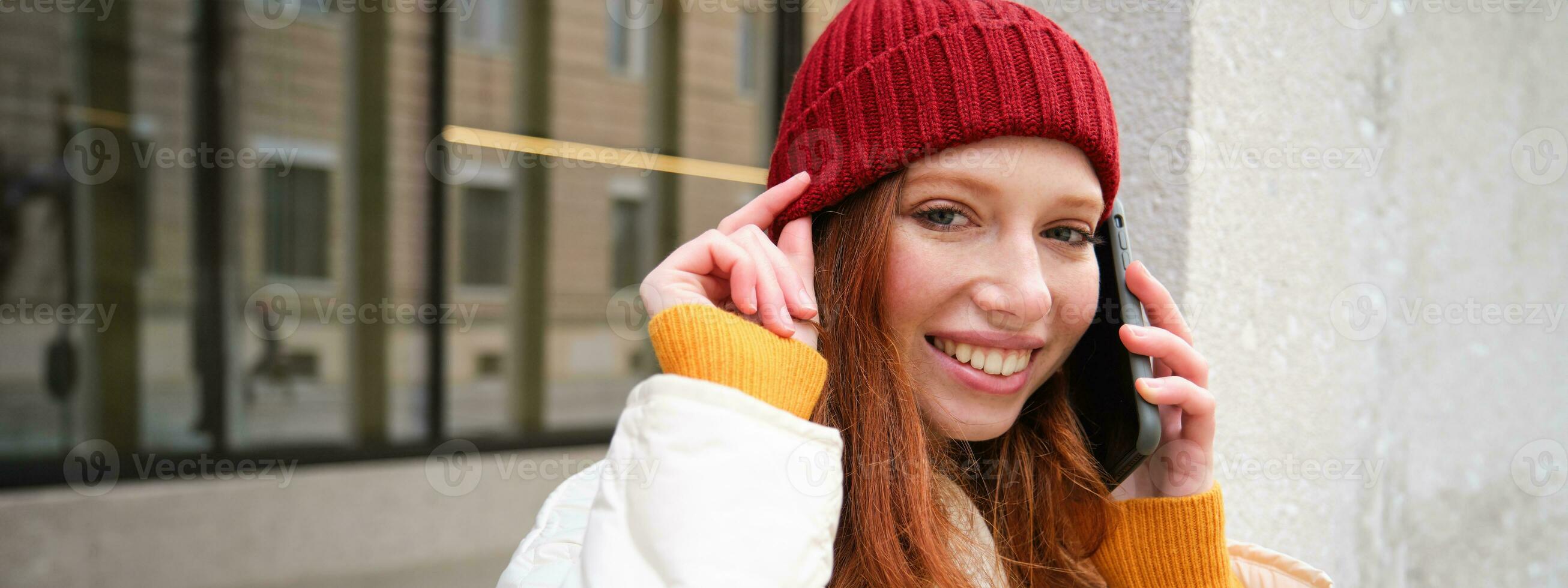 Young people and mobile connection. Happy redhead girl talks on phone, makes telephone call, stands outdoors with backpack and uses smartphone app photo