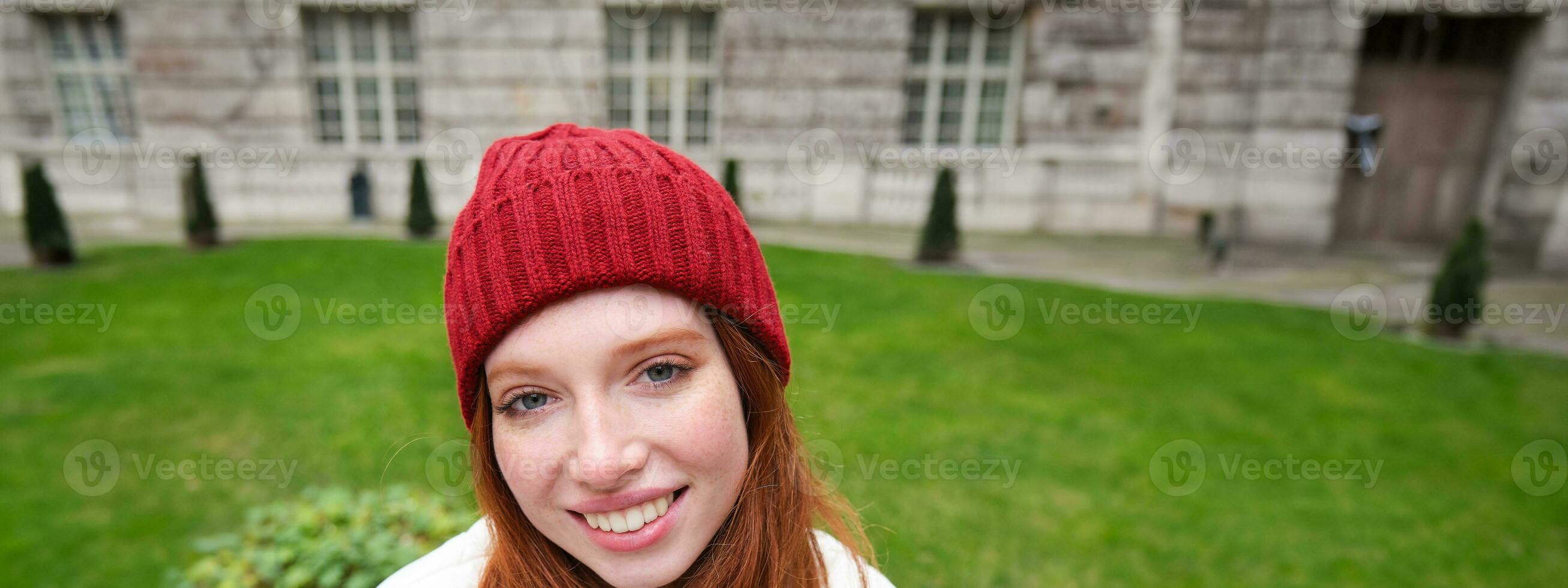 Vertical portrait of cute redhead female student in red hat and warm gloves, sits in park on bench, smiles and looks cute at camera photo